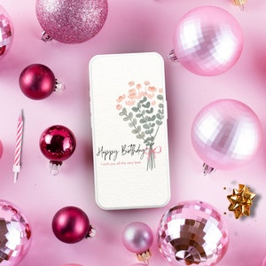 May include: A white phone case with a floral design and the text "Happy Birthday I wish you all the very best". The phone case is surrounded by pink ornaments on a pink background.