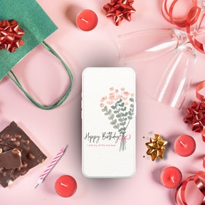 May include: A white smartphone with a pink and green floral design and the text "Happy Birthday I wish you all the very best" on the screen. The phone is surrounded by red and gold bows, candles, and chocolate.