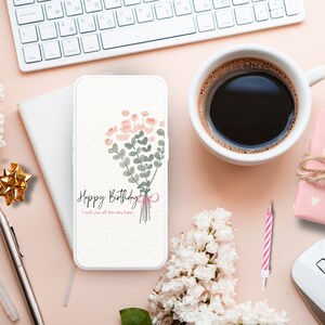 May include: A white smartphone with a floral design and the text "Happy Birthday I wish you all the very best" on the screen. The phone is on a pink surface with a white keyboard, a cup of coffee, a small gift box, a white mouse, and a pink and white striped candle.
