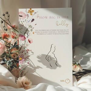 Op de afbeelding: Een witte kaart met een bloemenontwerp en de tekst "HOW BIG IS HER belly" in roze en gouden letters. De kaart bevat ook instructies voor een spel: "PULL THE RIBBON AND CUT WHERE YOU THINK WILL FIT AROUND THE MUMMY-TO-BE'S BELLY. THE PERSON WITH THE RIBBON CUT CLOSEST TO HER ACTUAL SIZE WINS!" en een silhouet van een zwangere vrouw.