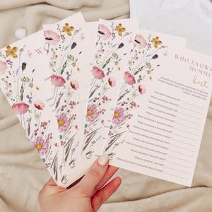 Op de afbeelding: Een set bloemen-thema "Wie kent mama het beste?" spelkaarten. De kaarten zijn roze met een bloemenpatroon en hebben vragen over de aanstaande moeder. De tekst op de kaarten is zwart.