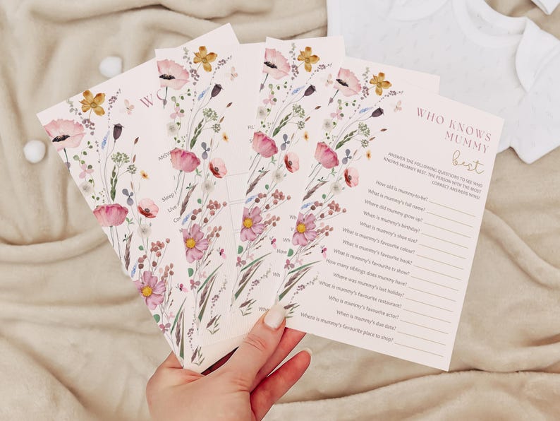 Op de afbeelding: Een set bloemen-thema "Wie kent mama het beste?" spelkaarten. De kaarten zijn roze met een bloemenpatroon en hebben vragen over de aanstaande moeder. De tekst op de kaarten is zwart.