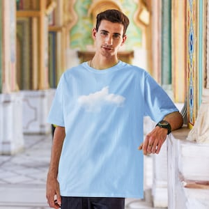 Puede incluir: Una camiseta azul claro con un estampado gráfico de nube blanca. La camiseta tiene cuello redondo y mangas cortas.