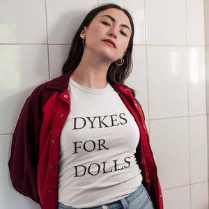 Puede incluir: Camiseta blanca con el texto negro "DYKES FOR DOLLS", combinada con una chaqueta roja y granate y vaqueros azul claro. La persona está apoyada contra una pared de azulejos blancos.