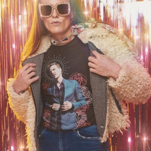 May include: A person wearing a black t-shirt with a graphic print of a man in a suit and a halo. The t-shirt is worn under a white fluffy jacket. The person is also wearing blue jeans and gold-rimmed sunglasses.