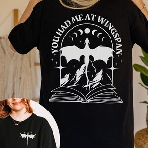 Peut inclure: T-shirt noir avec un motif blanc représentant un dragon, des montagnes et un livre ouvert. Le texte "YOU HAD ME AT WINGSPAN" est inscrit au-dessus du dragon. Une version plus petite du motif est sur une autre chemise.