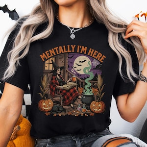 Może przedstawiać: Czarny t-shirt z krótkim rękawem z nadrukiem przedstawiającym czarownicę siedzącą na werandzie z książką, kubkiem, czarnym kotem, dyniami i kukurydzą. Tekst nad ilustracją brzmi MENTALLY I'M HERE, napisany pomarańczowymi wielkimi literami.