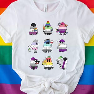 Op de afbeelding: Wit T-shirt met een ontwerp van verschillende gekleurde geesten die verschillende voorwerpen vasthouden. De geesten zijn allemaal in verschillende kleuren en hebben verschillende gezichtsuitdrukkingen.