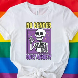 Peut inclure: T-shirt blanc avec un graphique violet et jaune d'un squelette tenant une tasse de café. Le graphique dit "NO GENDER ONLY ANXIETY".