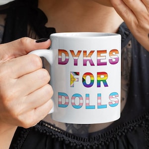 May include: White ceramic mug with the text "DYKES FOR DOLLS" in rainbow colors. The mug is held by a person with a tattoo on their hand that says "WORLD WEIRD GIRL".