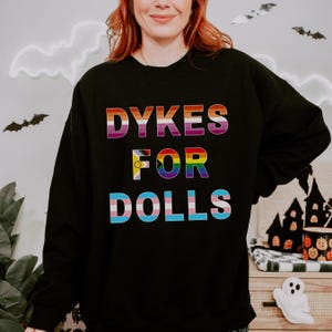 May include: Black sweatshirt with the text "DYKES FOR DOLLS" in rainbow colors. The text is layered with the colors of the lesbian, pride, and transgender flags. The sweatshirt is a casual, comfortable style.