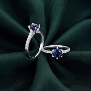 May include: Two silver-coloured rings featuring a round-cut dark blue gemstone set in a six-prong setting. The bands are crafted from a polished silver-toned metal and are accented with a row of small, clear, round-cut stones along the top half of the band.