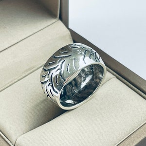 Puede incluir: Un anillo de plata con un diseño texturizado. El anillo está en una caja de anillo gris.