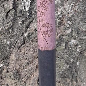 May include: A walking cane featuring a light purple upper shaft with a dark wood-burned vine and leaf design. The lower portion of the shaft is painted solid black, and the base is finished with a black rubberized non-slip ferrule tip.