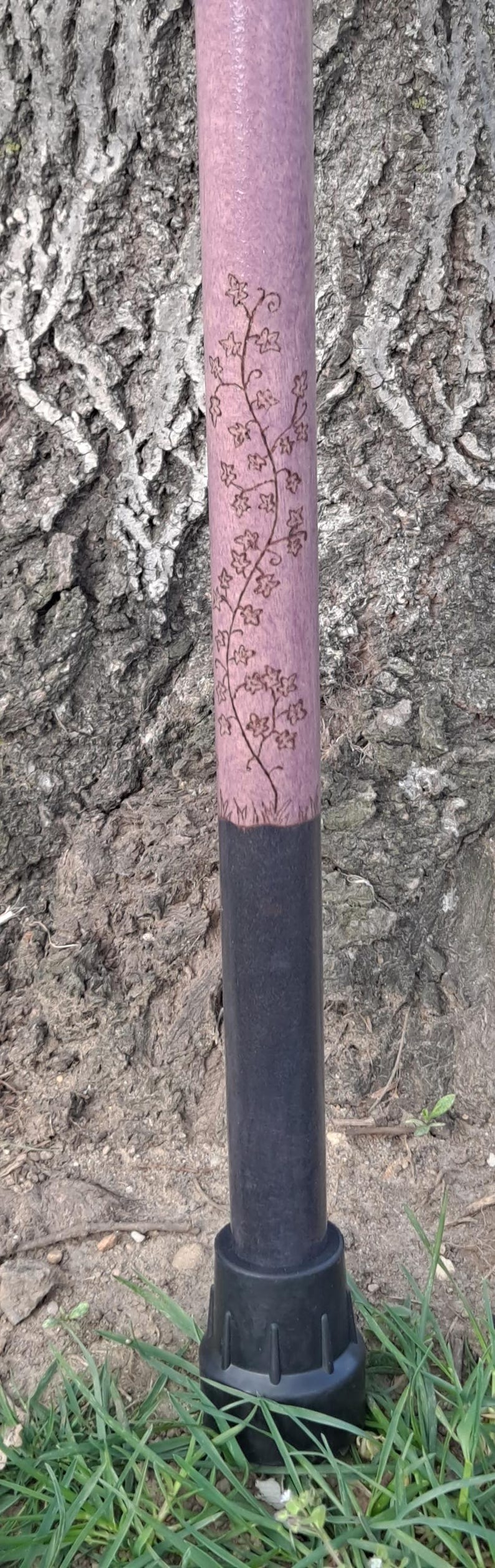 May include: A walking cane featuring a light purple upper shaft with a dark wood-burned vine and leaf design. The lower portion of the shaft is painted solid black, and the base is finished with a black rubberized non-slip ferrule tip.