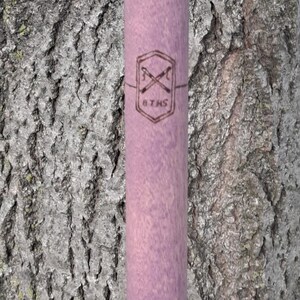 May include: A cylindrical lavender-colored wooden rod featuring a dark brown laser-engraved emblem. The emblem consists of a hexagon shape containing two crossed sticks and the text 8 THS printed clearly in the center of the wooden surface.