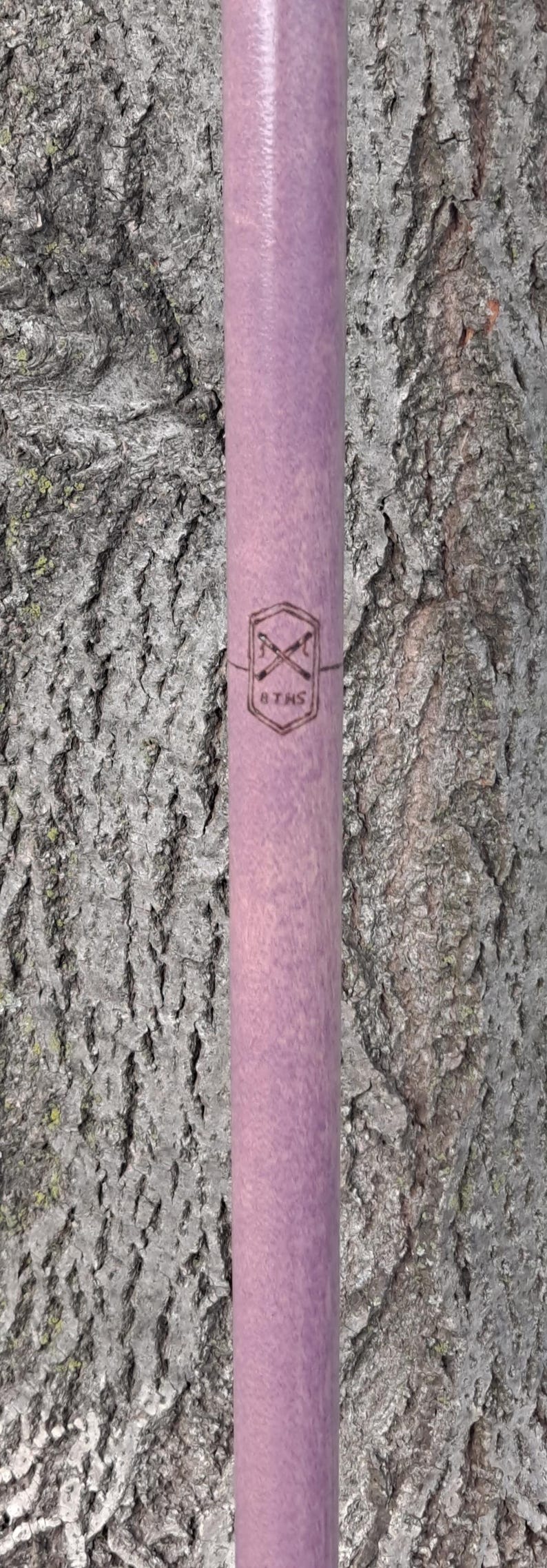 May include: A cylindrical lavender-colored wooden rod featuring a dark brown laser-engraved emblem. The emblem consists of a hexagon shape containing two crossed sticks and the text 8 THS printed clearly in the center of the wooden surface.