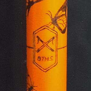 May include: A bright orange cylindrical object featuring black line art of butterflies and a central hexagonal emblem. The emblem contains a crossed pair of tools above the text 8THS, all rendered in a dark, etched-style graphic design.