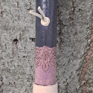 May include: A cylindrical wooden staff featuring a dark purple stained upper section with a small light-colored leather cord loop and a lower section finished in a lighter, natural beige tone. A floral design is etched into the wood transition.