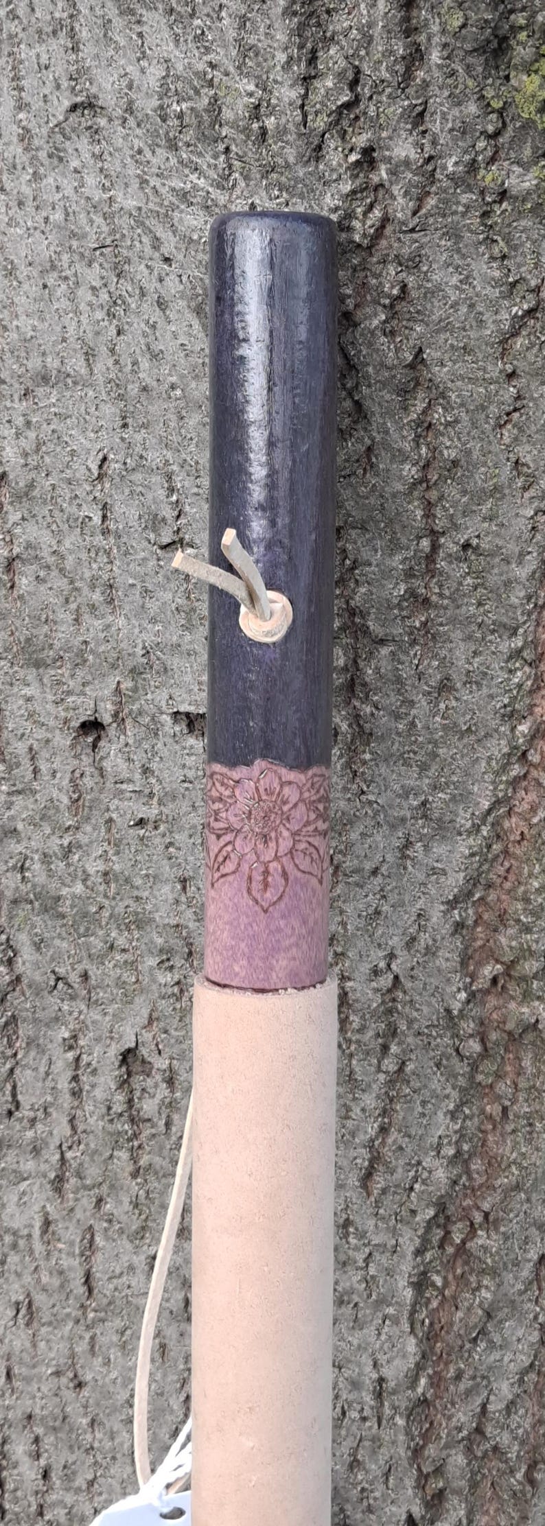 May include: A cylindrical wooden staff featuring a dark purple stained upper section with a small light-colored leather cord loop and a lower section finished in a lighter, natural beige tone. A floral design is etched into the wood transition.