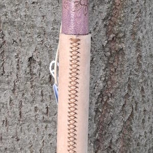May include: A wooden walking stick featuring a dark purple top section, a light purple middle section with floral engravings, and a light tan leather grip wrapped around the center with a decorative cross-stitch pattern and a white cord.