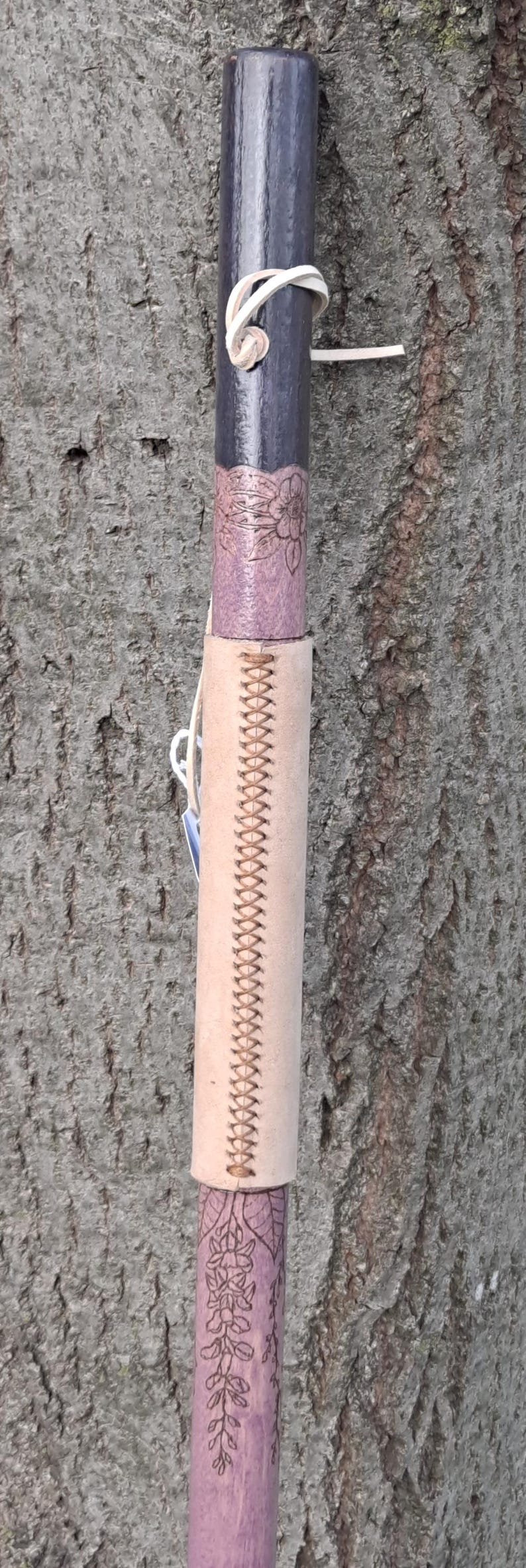 May include: A wooden walking stick featuring a dark purple top section, a light purple middle section with floral engravings, and a light tan leather grip wrapped around the center with a decorative cross-stitch pattern and a white cord.