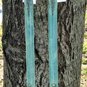 May include: Two long walking sticks with dark blue handles and light blue shafts featuring intricate floral engravings. Each stick includes a brown suede grip, a leather cord accent, and a small white tag attached near the handle area.