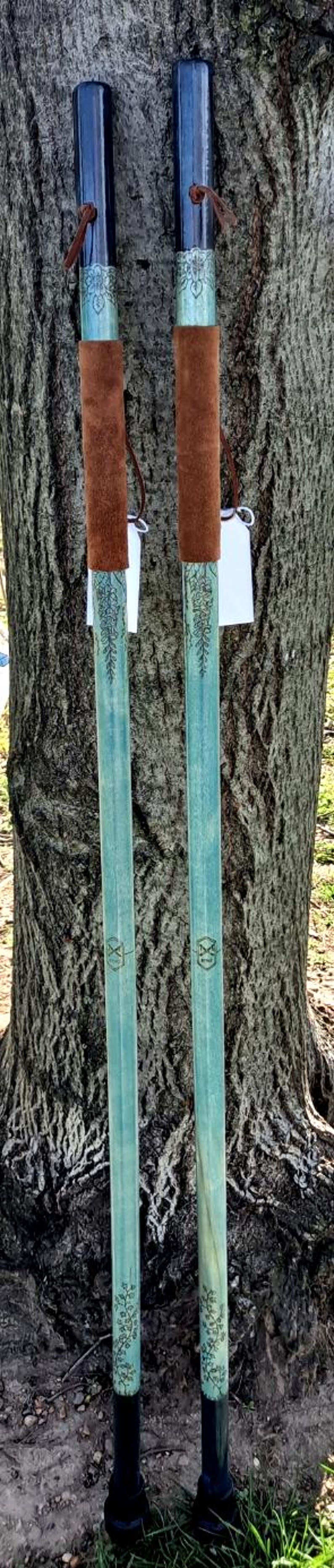 May include: Two long walking sticks with dark blue handles and light blue shafts featuring intricate floral engravings. Each stick includes a brown suede grip, a leather cord accent, and a small white tag attached near the handle area.