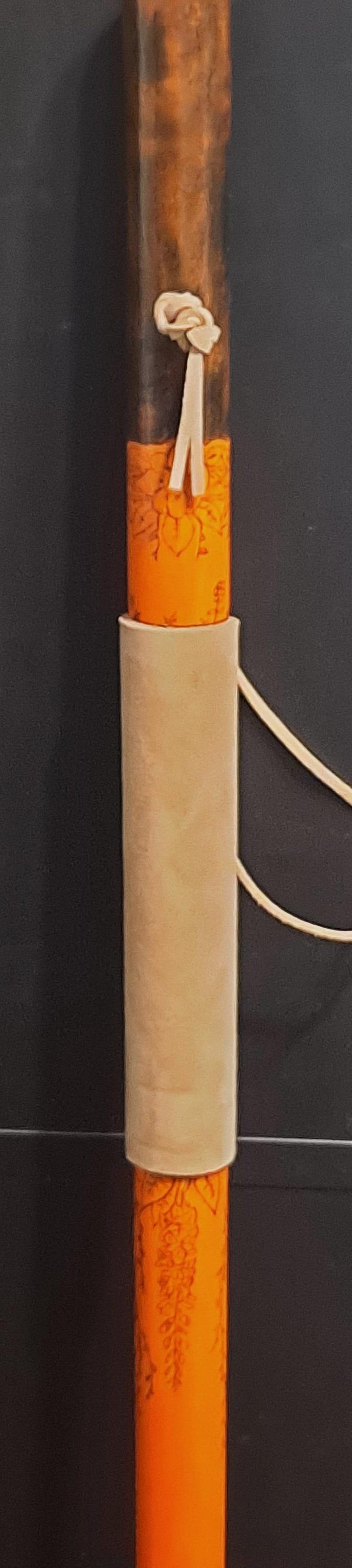 May include: A cylindrical walking stick featuring a dark brown wooden upper section, a bright orange middle section with etched floral patterns, and a smooth beige leather grip. A small white cord is tied in a knot near the top of the orange section.