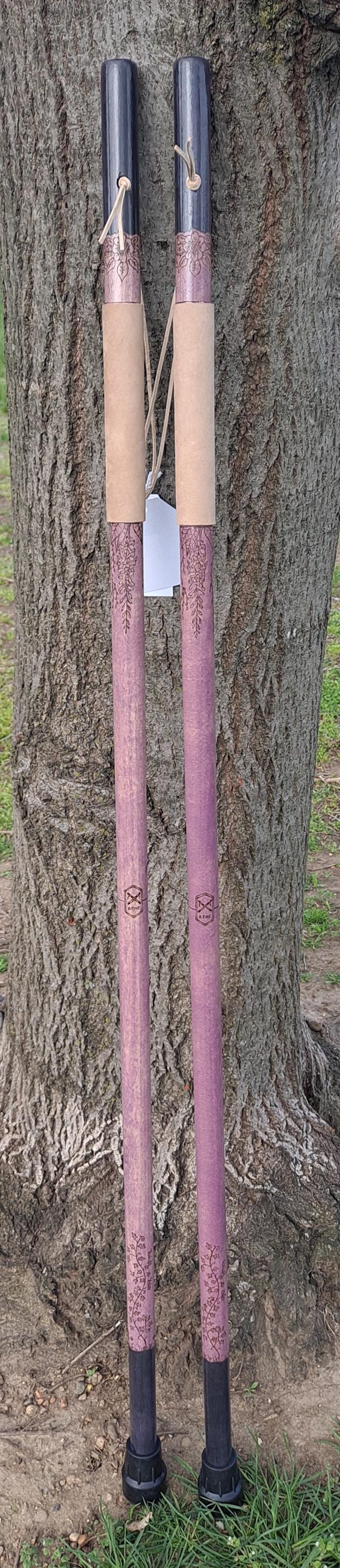 May include: Two matching walking sticks featuring a purple shaft with engraved floral patterns and a dark black handle. Each stick includes a tan leather grip, a small leather cord, and a black rubber tip at the base. A small logo is visible.