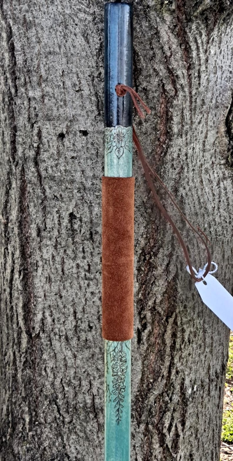 May include: A walking stick featuring a black top section, a light green middle section with a floral engraving, and a brown suede leather grip. A thin brown leather cord is tied near the top, and a small white blank tag hangs from the cord.