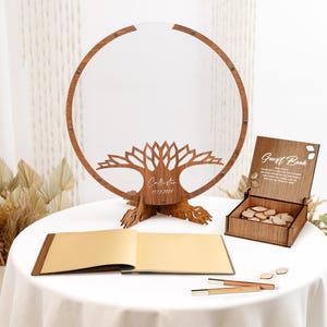 May include: A wooden guest book alternative with a tree design and a clear acrylic circle. The tree has a personalized inscription with the date "11.13.2021". The guest book is a wooden box with a lid and a sign that says "Guest Book". The box is filled with small wooden hearts. The guest book is on a white table cloth with a gold book and two pens.