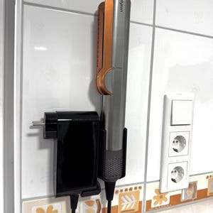 Könnte beinhalten: Ein silbernes und roségoldenes Haarstyling-Tool mit dem Wort "Dyson" auf der Seite, montiert an einer Wand mit einer schwarzen Steckdose und einer weißen Steckdose mit zwei Steckplätzen.