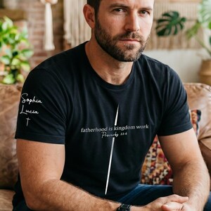 May include: Black t-shirt with the text "fatherhood is kingdom work Proverbs 22:6" and "Sophia Lucia" on the sleeve. The shirt is worn with blue jeans and a black watch. The person is sitting on a brown leather sofa.