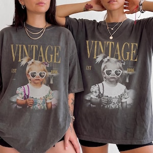 May include: A charcoal grey oversized t-shirt featuring a graphic print of a young child wearing heart-shaped sunglasses. The text VINTAGE EST 1996 is printed in a distressed gold font above and beside the child. The shirt has a crew neckline.