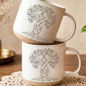May include: Two stacked ceramic mugs with a speckled off-white finish and a tan base. Each mug features a gray line-art illustration of a tree with intertwined trunks, a heart-shaped canopy, and the names Amelia and Jacob with the date 08.15.26.