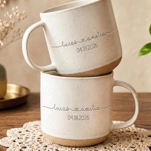 May include: Two stacked ceramic mugs with a speckled off-white glaze and a natural clay base. Each mug features the printed text "lucas & amelia" with a small heart icon between the names and the date "04.18.2026" printed directly underneath.