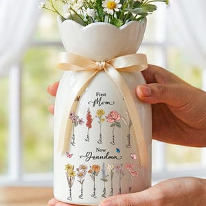 May include: A white ceramic vase with a cream ribbon, filled with white and yellow flowers. The vase features floral illustrations and the text "First Mom, Now Grandma" with names. A decorative item for the home.