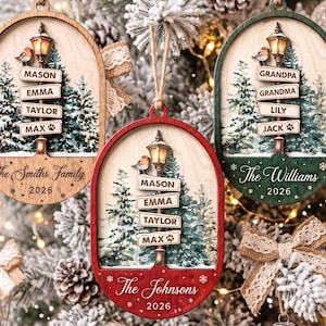 May include: Three oval wooden Christmas ornaments featuring a street lamp with a bird, snow-covered pine trees, and personalised name signs. The ornaments are coloured in natural wood, red, and green, each displaying family names and the year 2026.