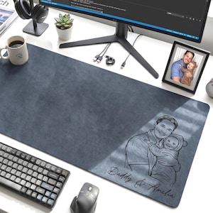 May include: A dark blue rectangular desk mat with a personalized sketch of a father and child, with the text "Daddy & Amelia". A keyboard, mouse, and coffee mug are on the white desk.