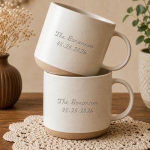 May include: Two off-white ceramic mugs stacked vertically, each featuring a smooth handle and a natural clay-colored base. The text "The Bonannos" and "05.20.2026" is engraved in a cursive font on the front of both mugs, which have a speckled finish.