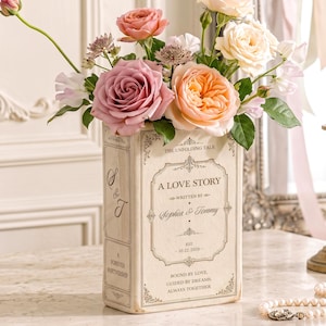 May include: A rectangular cream-colored vase shaped like a book, featuring the text A LOVE STORY, WRITTEN BY Sophia & Tommy, EST. 10.22.2026, and BOUND BY LOVE, GUIDED BY DREAMS, ALWAYS TOGETHER. It holds a bouquet of pink, peach, and white roses.