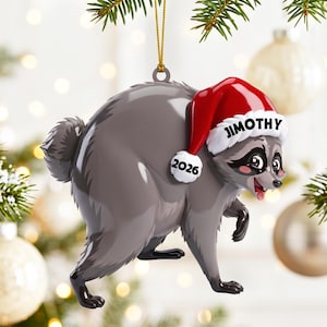 Peut inclure: Une d&eacute;coration de No&euml;l en forme de raton laveur gris portant un bonnet de P&egrave;re No&euml;l rouge &agrave; bord blanc. Le bonnet porte le nom JIMOTHY en noir et l'ann&eacute;e 2026 sur le pompon blanc. Le raton laveur a des taches noires autour des yeux.