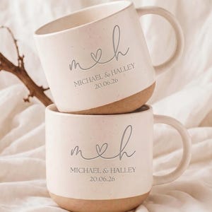 May include: Two stacked ceramic mugs with a speckled cream finish and a natural tan base. Each mug features the gray printed text "m ♡ h" above the names "MICHAEL & HALLEY" and the date "20.06.26" in a clean, sans-serif font style.