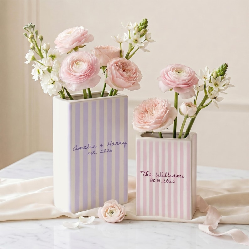 May include: Two rectangular ceramic vases with vertical stripes in light purple and light pink. The purple vase features the text Amelia + Harry est. 2026, while the pink vase displays The Williams 08.18.2026. Both contain arrangements of pink ranunculus and white flowers.
