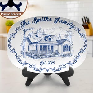 May include: Oval white ceramic platter with a blue illustration of a house, mountains, and text that reads "The Smiths Family" and "Est. 2025". The platter is displayed on a black plastic stand.