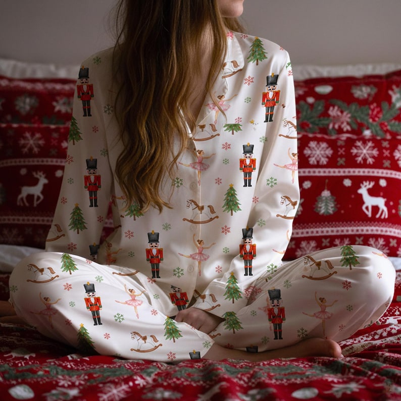 May include: Cream-colored pajama set featuring a festive print of nutcrackers, ballerinas, Christmas trees, and rocking horses. The long-sleeved top and pants are set against a red patterned background, creating a cozy holiday theme.
