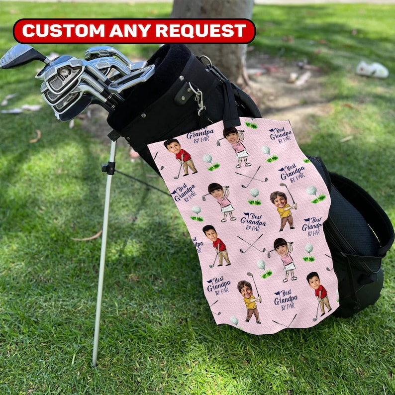 May include: A black golf bag with golf clubs and a pink golf towel. The towel has a repeating pattern of cartoon figures, golf balls, and crossed golf clubs. Text on the towel reads "Best Grandpa by Par."