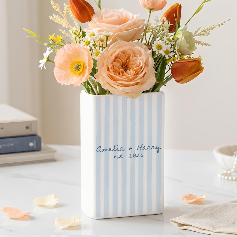 May include: A rectangular white ceramic vase featuring vertical light blue stripes. The front is printed with the text Amelia + Harry est. 2026 in dark blue script. The vase is filled with a bouquet of peach, orange, and white artificial flowers.