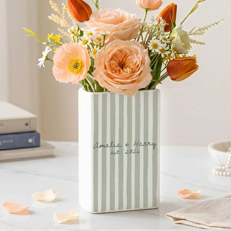 May include: A rectangular ceramic vase featuring vertical sage green stripes on a white background. The front is personalized with the text Amelia + Harry est. 2026 in a dark green script font. The vase is filled with a bouquet of peach roses.